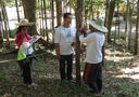 Four people measuring a tree. They are wearing long pants and white long-sleeve shirts. Three people clustered around a thin tall tree with a pink measuring tape around it. The fourth person is writing on a clipboard. One of the people around the tree is holding a yellow measuring tape connecting to something out of the image.
