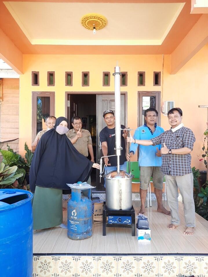 A woman with face mask and burka and 5 men in informal clothes posing next to distillation equipment in and outdoor patio. Behind them there is the entrance of a building.
